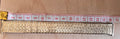 A close-up of a gold-toned metal link bracelet or chain scale against a light wood background, with a red-and-white measuring tape laid above it for size reference.