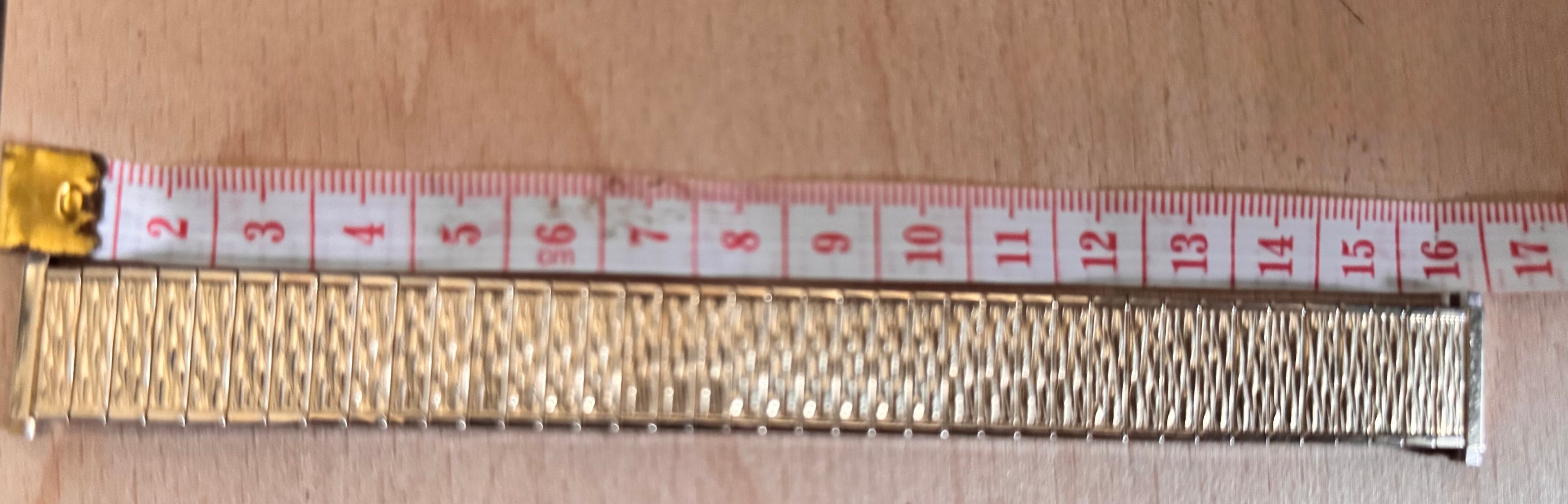 A close-up of a gold-toned metal link bracelet or chain scale against a light wood background, with a red-and-white measuring tape laid above it for size reference.