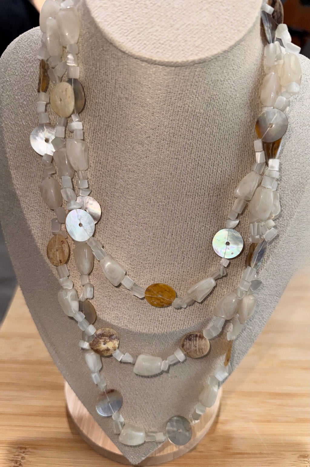 A multi-strand beaded necklace displayed on a beige fabric jewelry bust. The necklace features a mix of natural materials including disc shells, flat beads, and small white beads in neutral tones of beige, gray, and brown.