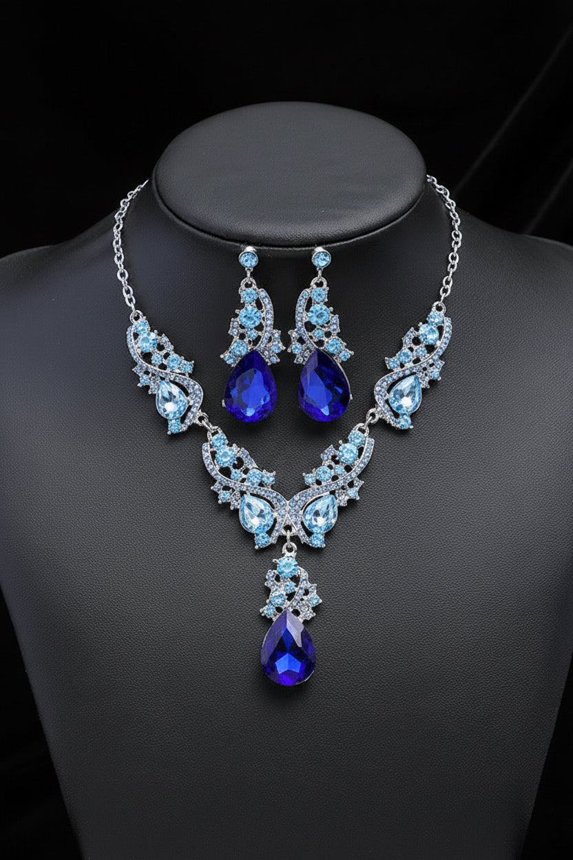 A two-piece jewelry set featuring a silver-tone necklace, matching drop earrings, and a teardrop pendant, all adorned with sapphire colour faux gems surrounded by a light blue faux gems in swirls. The necklace and earrings are set on a Charcoal bust. Please note all the larger gems are Sapphire colour blue